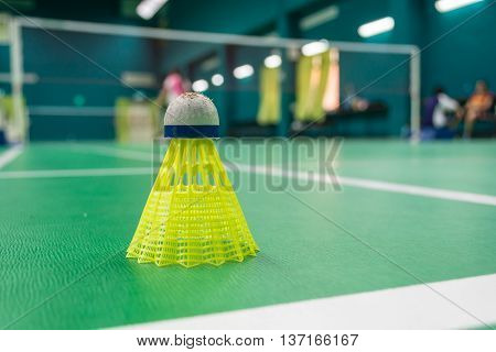 Yellow plastic badminton shuttlecock on a green floor with blurred players in badminton court.