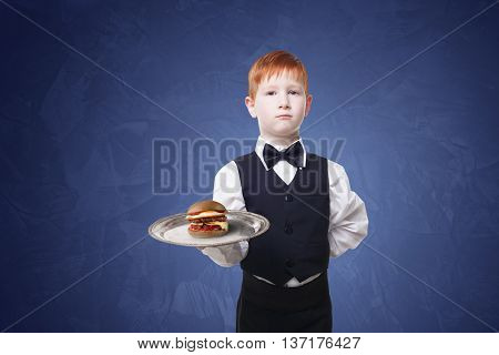 Little waiter stands with tray serving hamburger. Redhead child boy in suit plays restaurant servant, gives burger at blue background