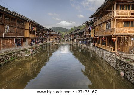Zhaoxing Dong Village Guizhou Province China - April 8 2010: Rural River with cobblestone seafront promenade and wooden houses with trade rows on both shores village ethnic minorities sunny day