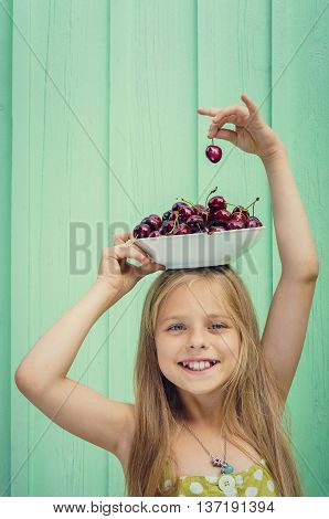 Beautiful blond girl on a background of turquoise wall holding a plate with cherries on her head. Space for text
