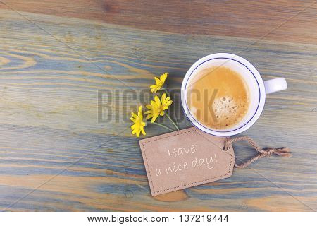 Coffee cup and daisy flowers with wish cardboard label on wooden table. Have a nice day romantic message. View from above