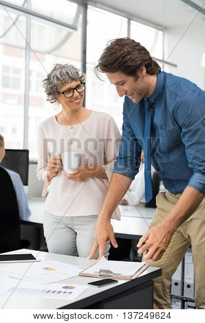 Senior businesswoman looking at young business man. Mature business woman holding a cup of tea and working with young businessman. Assistant and senior leader in a conversation in a modernoffice.
