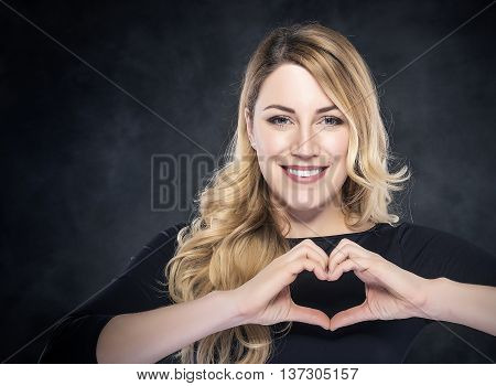 Portrait of beautiful young blond woman showing heart sign over dark background.