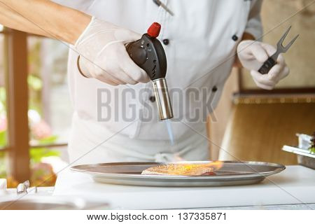 Man's hand with torch burner. Torch blowing fire on meat. Chef is preparing duck breast. Be careful not to overcook.