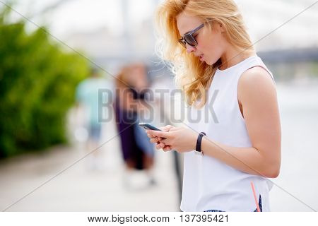 Casual blone girl in sunglasses standing on city street and using mobile phone
