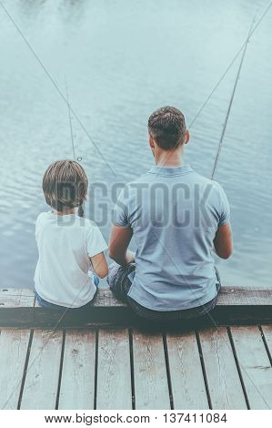 Enjoying day fishing. Rear view of father and son fishing while sitting on quayside together
