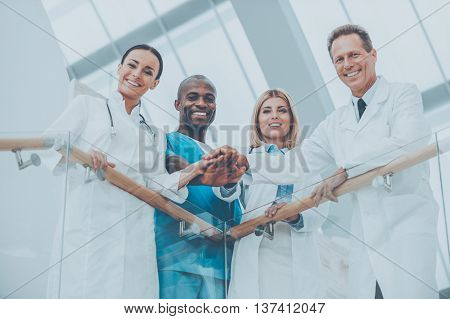 Confident medical experts. Low angle view of four happy medical doctors standing close to each other and holding their hands together while leaning at the handrail