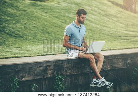 Surfing web outdoors. Handsome young man in polo shirt working on laptop while sitting on quayside