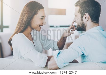 Enjoying every minute together. Playful young loving couple sitting together on the couch and looking at each other with smile while woman touching nose of her boyfriend