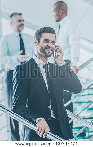 Sharing great news. Low angle view of confident young man in formalwear talking on the mobile phone and smiling while moving down by staircase with people in the background