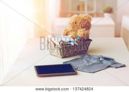 babyhood, motherhood, clothing, technology and object concept - close up of baby clothes and toys for newborn boy in basket with tablet pc computer at home