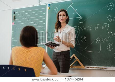 Teacher or docent or educator giving while lesson in front of a blackboard or board a sheet of paper and educate or teaching students or pupils or mates in a school or class.