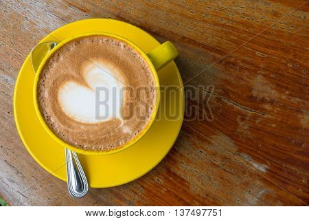 A cup of coffee with heart pattern in a yellow cup on wooden background