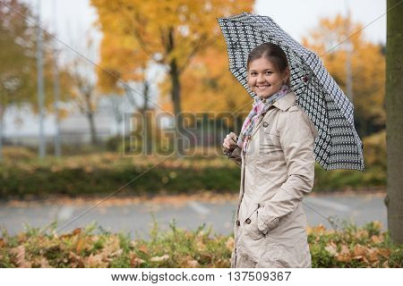 Young woman standing under umbrella in autumn park wearing beige mackintosh