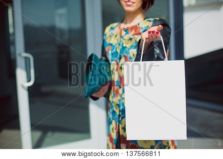 retail shopping concept. Woman holding a white paper shopping bag with space for text. Selective focus foreground package. Sunny day happy shopping.