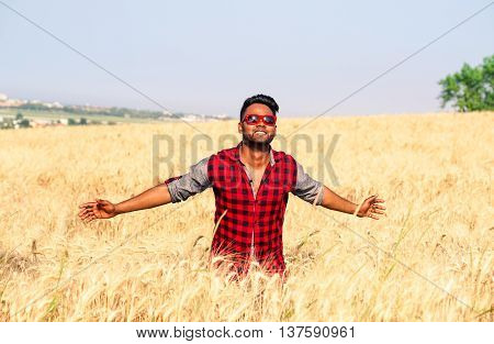Young indian man walking in wheat field - African american guy open arms at sky through a corn field - Asian farmer with open arms towards the sun - Concept of freedom and joy for successful achieved