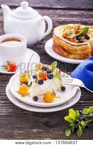 Homemade breakfast. Fresh cheesecake with Nordic berries cloudberries and blueberries in plain wooden background served with cloudberry tea in a white ceramic bowl. Decorated branches forest blueberries