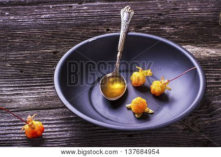 Black simple dish with a spoon Arctic cloudberry seed oil full with ripe fresh berries north cloudberries in plain wooden background in rustic background.