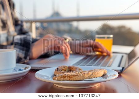 Working Breakfast Person Working on Laptop Computer at Cafe Terrace on Table with Coffee Mug Orange Juice and Bakery Cake Asian Urban Landscape on Background