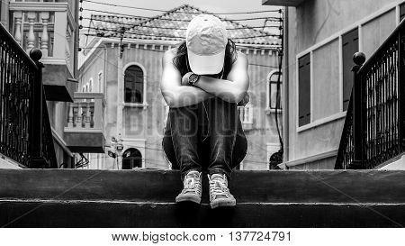 sad woman wearing a hat and hugging knees on stair backand white