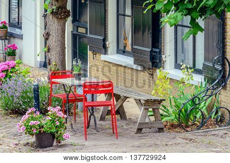 Simple bright red chairs with a small table and an old wooden bench draw attention of passers in a historic Dutch village with many historic buildings.