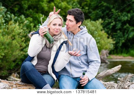 Happy lovers in vacation sitting at waterside on a trunk clinking beer bottles