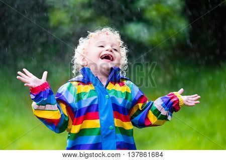 Little blond curly boy in a waterproof jacket in rainbow color playing in the rain. Kids having fun outdoors in autumn shower.