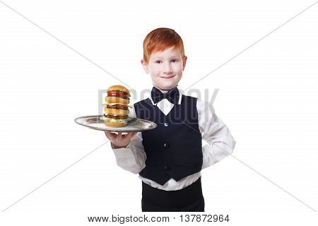 Little waiter stands with tray serving big double hamburger. Smiling redhead child boy in suit plays restaurant servant, gives burger isolated at white background