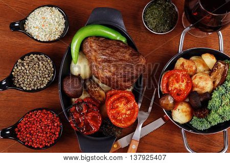 fresh roast beef meat rib eye steak with tomatoes,potatoes, hot chili pepper gern and pink peppercorn saucer, cutlery, set of spice, and red wine glass on light walnut table