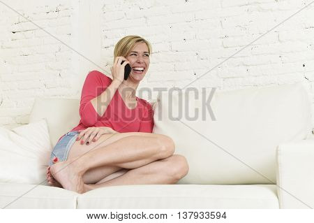 young beautiful Caucasian woman with shorts and modern hair style happy on couch talking on mobile phone relaxed and cheerful laughing and smiling at home living room in communication concept