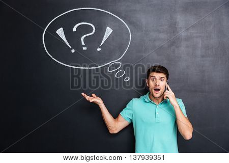Amaized handsome young man talking on mobile phone over blackboard background with blank speech bubble