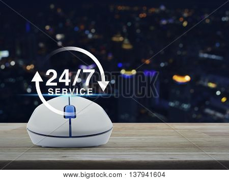 Wireless computer mouse with button 24 hours service icon on wooden table in front of blurred light city tower Full time service concept