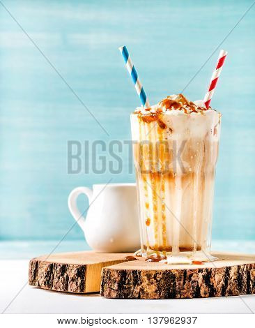Latte macchiato with whipped cream and caramel sauce in tall glass with two straws on wooden board over blue painted wall background, selective focus, copy space, vertical composition