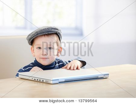 Adorable niño pequeño jengibre con tapa, jugando con la computadora portátil en la mesa.