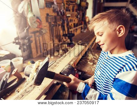 carpentry, woodwork, building and people concept - happy little boy with hammer hammering wood plank at workshop