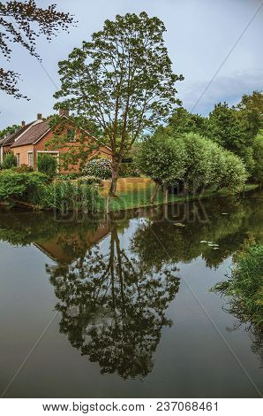 Charming Small Canal Next To Rustic House With Lush Garden And Trees Reflected On The Water In Cloud