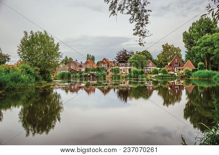 Charming Wide Canal Next To Rustic Houses With Green Bushes And Trees Reflected On The Water In Clou