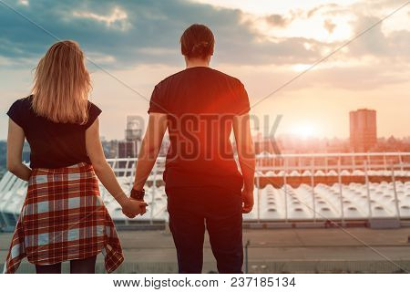 A Boy With A Girl Taking Hands On His Hands Watching The Sunset In The Evening City. Silhouette Of L