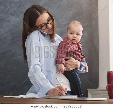 Happy Mother Working At Office With Her Baby. Young Woman Talking On Phone And Writing Notes While S