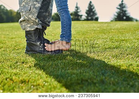 Close Up Feet Of Soldier And Daughter. Leather Military Boots And Naked Feet On The Meadow.