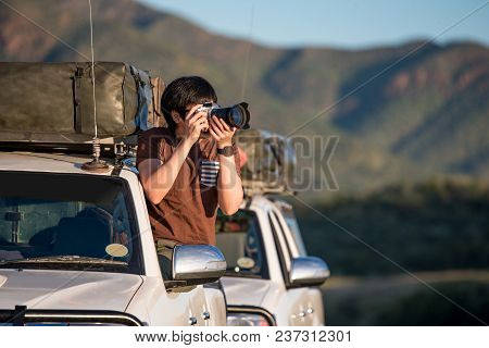 Young Asian Man Traveler And Photographer Sitting On The Car Window Photographing Sunrise During Roa