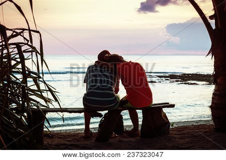 A Young Couple Sits On Bench Under A Palm Tree And Looks At The Sunset On The Sea. Honeymoon Couple 