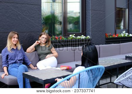 Young Female Friends Talking At Cafe And Sitting On Sofa Near Window With Flowers.. Concept Of Frien