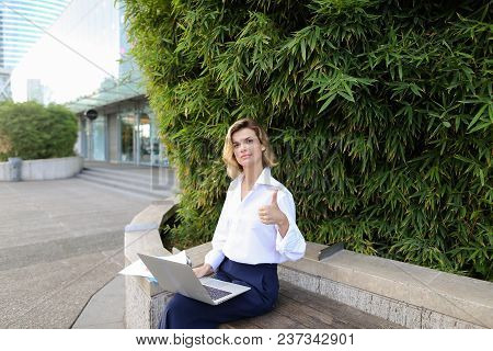 Caucasian Businesswoman Working With Laptop And Papers In Open Air Near Green Plant, Showing Thumbs 