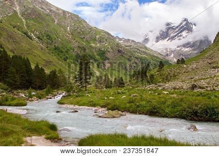 Colorful View Of Green And Snowy Mountains With Elbrus Summit Under Fluffy White Clouds.