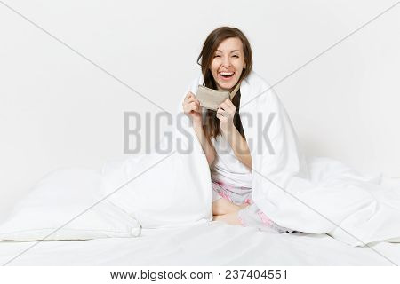 Fun Tired Young Woman Sitting In Bed With Sleep Mask, Sheet, Pillow, Wrapping In Blanket Isolated On