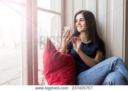 Sexy Brunette Girl Wears Blue T-shirt Holds Smartphone Sitting On Wide Windowsill With Big Red Pillo