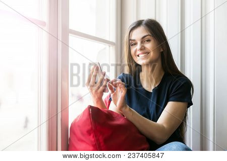 Sexy Brunette Girl Wears Blue T-shirt Holds Smartphone Sitting On Wide Windowsill With Big Red Pillo