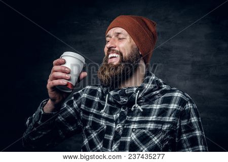 Brutal Bearded Male Dressed In A Hat And Fleece Shirt, Drinks Coffee From A Paper Glass.