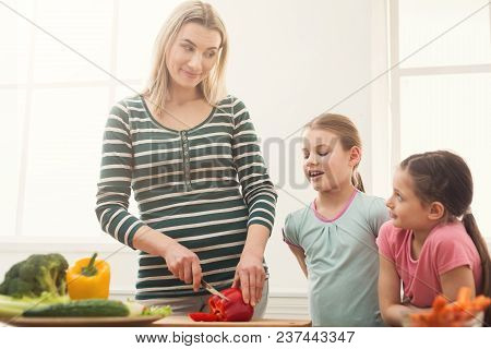 Beautiful Woman With Her Daughters Looks Happy While Cooking Salad In The Kitchen Over Window. Healt